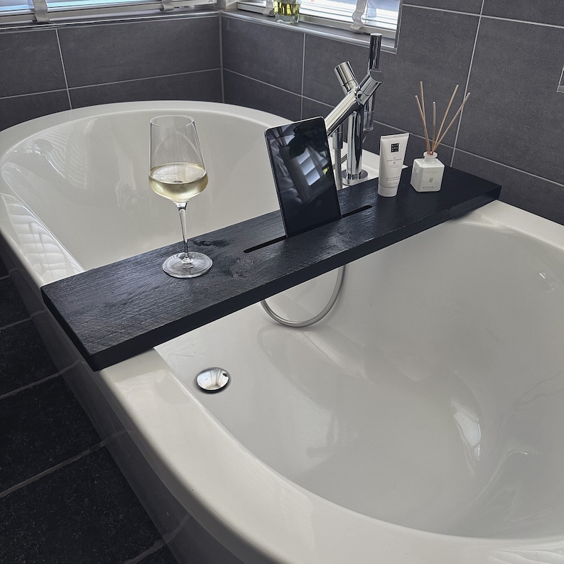 May include: A black wooden bath tray with a glass of white wine, a tablet, and toiletries. The tray is placed on a white bathtub.