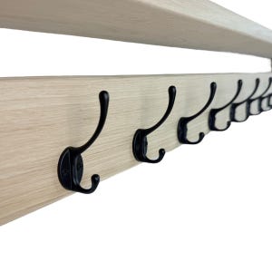 Puede incluir: Un perchero de madera clara con m&uacute;ltiples ganchos de metal negro. Los ganchos est&aacute;n espaciados uniformemente a lo largo del perchero, que tambi&eacute;n cuenta con una repisa en la parte superior. El dise&ntilde;o es simple y funcional.
