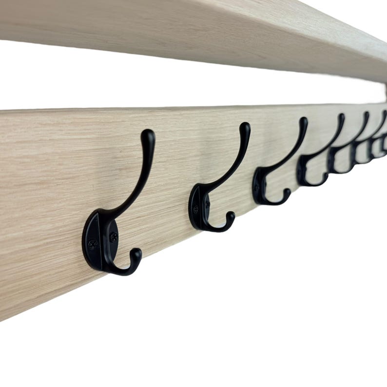 Puede incluir: Un perchero de madera clara con m&uacute;ltiples ganchos de metal negro. Los ganchos est&aacute;n espaciados uniformemente a lo largo del perchero, que tambi&eacute;n cuenta con una repisa en la parte superior. El dise&ntilde;o es simple y funcional.