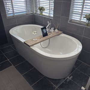 Puede incluir: Una ba&ntilde;era blanca con una bandeja de madera para productos de ba&ntilde;o. La bandeja tiene una copa de vino, una tableta y algunas botellas de productos de ba&ntilde;o. La ba&ntilde;era est&aacute; en un ba&ntilde;o con paredes de azulejos grises y una ventana.