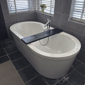 May include: A white bathtub with a black wooden bath tray. The bathtub is in a bathroom with gray tiled walls.