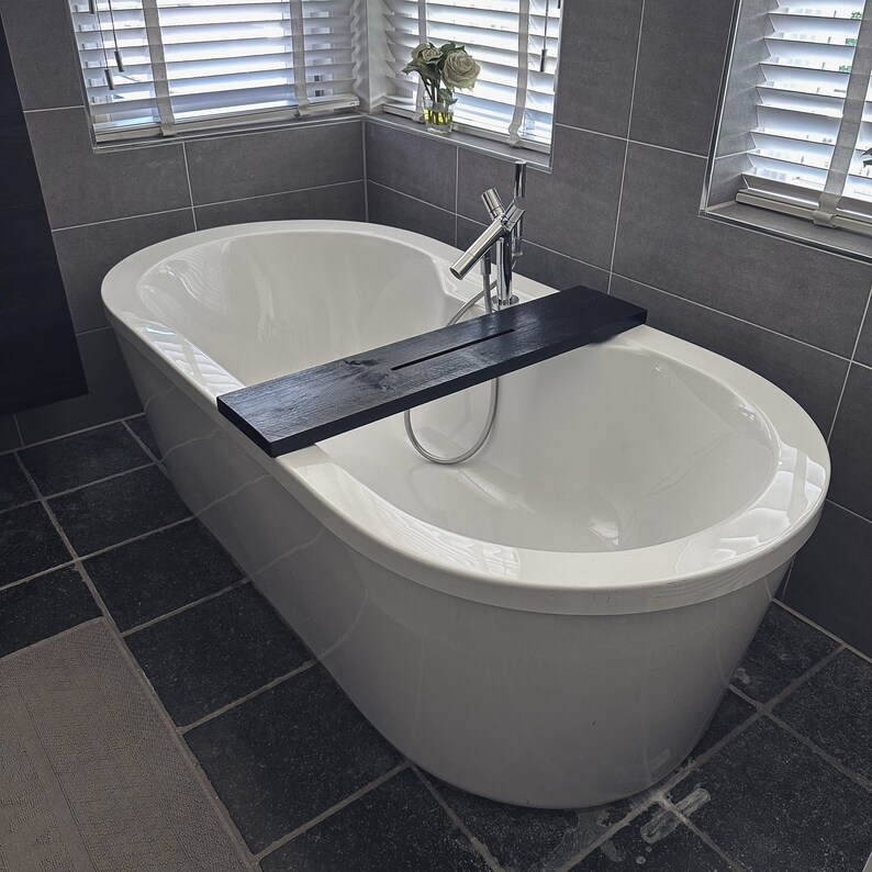 May include: A white bathtub with a black wooden bath tray. The bathtub is in a bathroom with gray tiled walls.