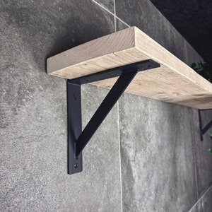 Op de afbeelding: Zwarte metalen plankdragers met een houten plank. De dragers zijn schuin en hebben een eenvoudig ontwerp.