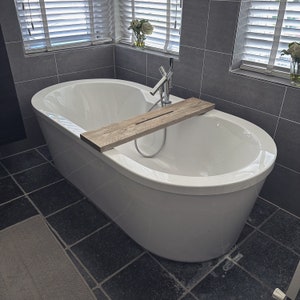 Puede incluir: Una ba&ntilde;era ovalada blanca con un estante de madera en la parte superior. La ba&ntilde;era est&aacute; en un ba&ntilde;o con paredes de azulejos grises y un suelo de azulejos negros.