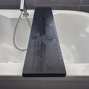 May include: Black wooden bath tray with a cutout for a glass of wine or a book. The tray is placed on the edge of a white bathtub.