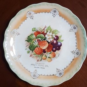 Peut inclure: Une assiette en céramique blanche avec un bord vert et orange. Au centre de l'assiette, une image peinte de fraises, de raisins et de fleurs. Le texte "Compliments of Argys Merc. Co., Salida, Colo." est imprimé en or sur l'assiette.