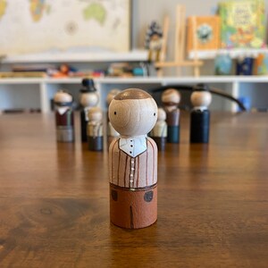 May include: A wooden peg doll with a brown head, a white shirt with brown stripes, and brown pants. The doll is standing on a wooden table.
