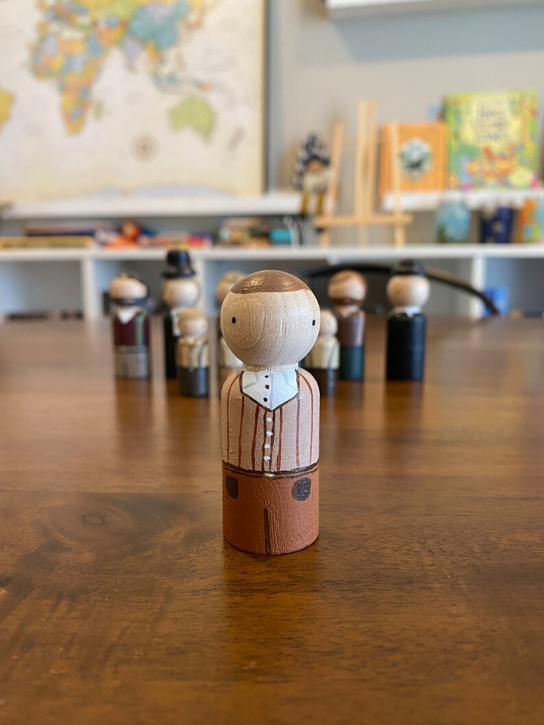 May include: A wooden peg doll with a brown head, a white shirt with brown stripes, and brown pants. The doll is standing on a wooden table.