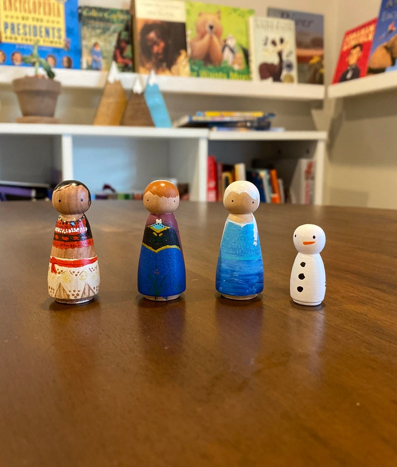 K&ouml;nnte beinhalten: Vier Holz-Steckpuppen, bemalt, um Figuren aus beliebten Animationsfilmen zu &auml;hneln. Die Puppen stehen auf einer Holzoberfl&auml;che. Die Puppen sind in verschiedenen Farben bemalt, darunter Blau, Rot, Wei&szlig; und Schwarz.