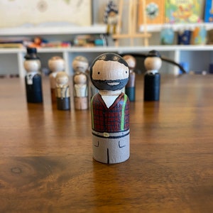 May include: A hand-painted wooden peg doll with a black beard, red and black plaid shirt, green suspenders, and gray pants.