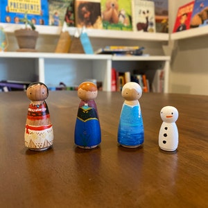 K&ouml;nnte beinhalten: Vier handbemalte Holz-Steckpuppen, die an Figuren aus beliebten Animationsfilmen erinnern. Die Puppen stehen auf einer Holzoberfl&auml;che. Die Puppen sind in verschiedenen Farben bemalt, darunter Rot, Blau, Wei&szlig; und Schwarz. Die Puppen sind etwa 10 cm gro&szlig;.