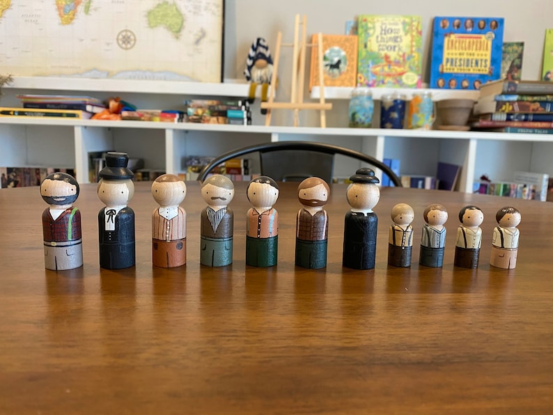 May include: A set of 11 wooden peg dolls, each painted to resemble a person with different facial features, clothing, and hairstyles. The dolls are standing on a wooden table.