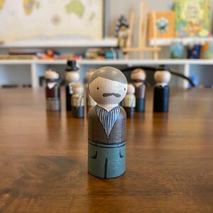 May include: A hand-painted wooden peg doll with a brown shirt, green pants, and a gray mustache. The doll is standing on a wooden surface.