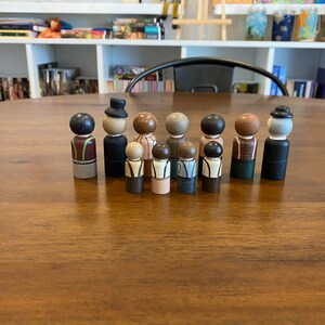 May include: A set of 10 wooden peg dolls, painted in various colors and designs. The dolls are all standing in a row on a wooden table. The dolls are all wearing different clothes and have different hairstyles.