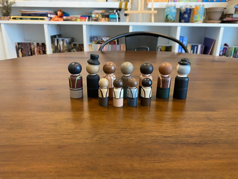 May include: A set of 10 wooden peg dolls, painted in various colors and designs. The dolls are all standing in a row on a wooden table. The dolls are all wearing different clothes and have different hairstyles.