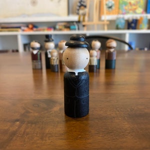 May include: A wooden figurine of a person wearing a black hat and a black suit. The figurine has a white face with black eyes and a white collar.