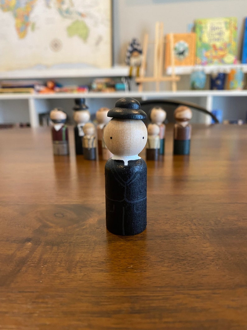 May include: A wooden figurine of a person wearing a black hat and a black suit. The figurine has a white face with black eyes and a white collar.