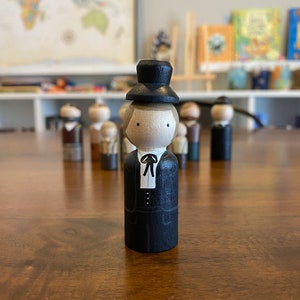 May include: A wooden peg doll wearing a black suit and a black top hat. The doll has a painted face with brown hair and is standing on a wooden surface.