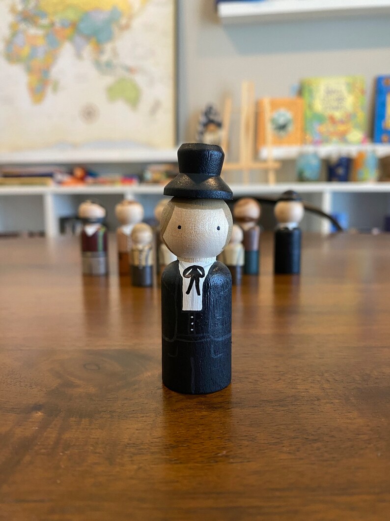 May include: A wooden peg doll wearing a black suit and a black top hat. The doll has a painted face with brown hair and is standing on a wooden surface.