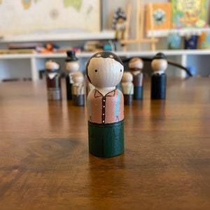 May include: A hand-painted wooden peg doll with black hair, a brown shirt, blue suspenders, and green pants.