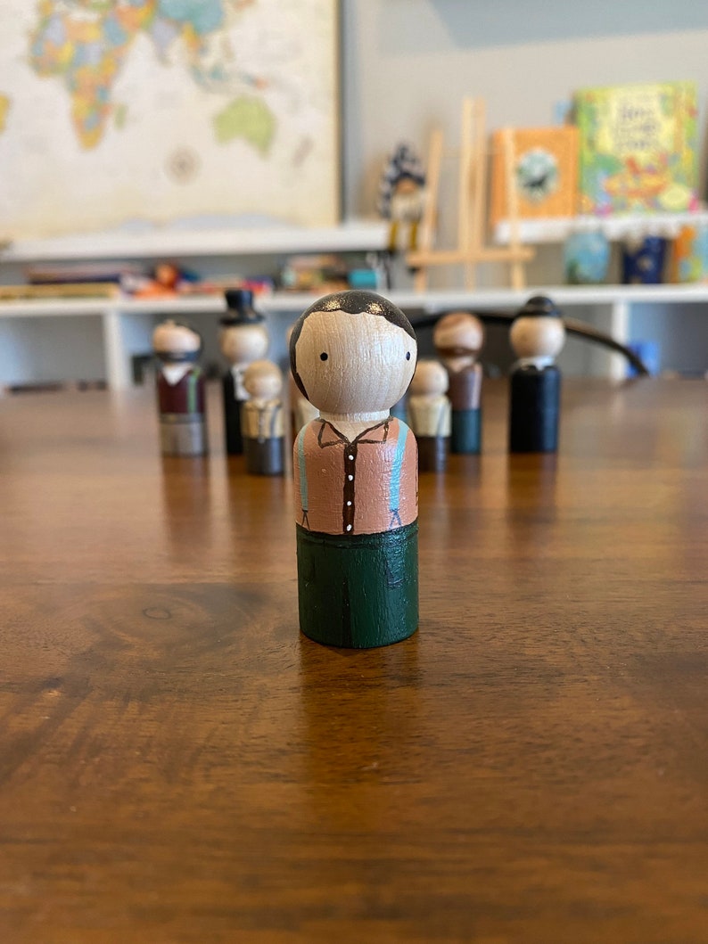 May include: A hand-painted wooden peg doll with black hair, a brown shirt, blue suspenders, and green pants.