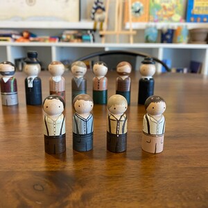 May include: A group of 11 wooden peg dolls, each painted to resemble a person wearing different clothing. The dolls are standing on a wooden table.
