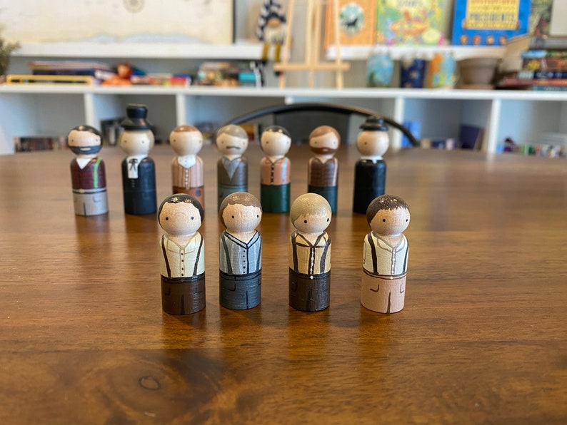 May include: A group of 11 wooden peg dolls, each painted to resemble a person wearing different clothing. The dolls are standing on a wooden table.