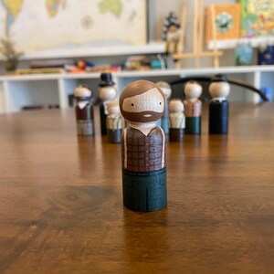 May include: A wooden peg doll with a brown beard and plaid shirt, green pants, and a brown hat. The doll is standing on a wooden table.