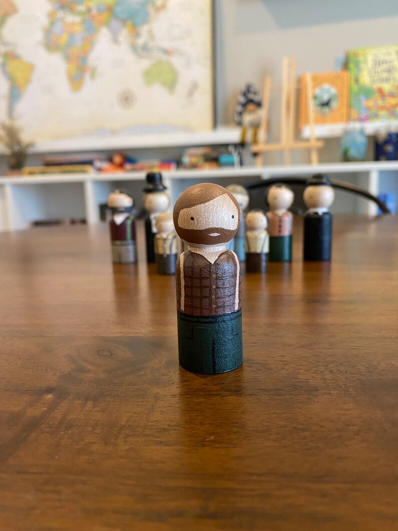 May include: A wooden peg doll with a brown beard and plaid shirt, green pants, and a brown hat. The doll is standing on a wooden table.