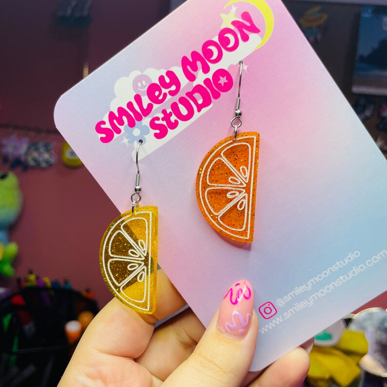May include: A pair of orange and yellow glitter lemon slice earrings with white outlines. The earrings are on a pink card with the text "Smiley Moon Studio" in white.