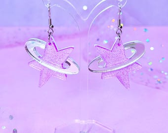 May include: A pair of dangling earrings featuring a five-pointed star shape with a translucent purple glitter finish. Each star is encircled by a clear, smooth ring, creating a planetary effect. The earrings hang from silver-toned metal hooks.