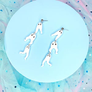 Peut inclure: Une paire de boucles d'oreilles pendantes en forme de fantôme blanc avec des crochets argentés. Chaque boucle d'oreille présente trois fantômes empilés les uns sur les autres.