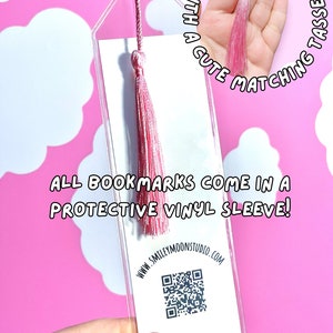 Op de afbeelding: Een transparante acryl bladwijzer met een roze kwast. De bladwijzer heeft een witte achtergrond met een QR-code en de tekst "WWW.SMILEYMOONSTUDIO.COM". De tekst "ALL BOOKMARKS COME IN A PROTECTIVE VINYL SLEEVE!" is op de bladwijzer gedrukt. De bladwijzer wordt in een hand met roze nagellak gehouden.