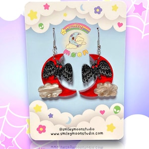 Puede incluir: Pendientes de media luna roja con una silueta de murci&eacute;lago negro y un detalle de nube blanca. Los pendientes est&aacute;n sobre un fondo azul y blanco con estrellas y el texto "@smileymoonstudio www.smileymoonstudio.com".