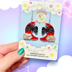 Puede incluir: Un par de pendientes de media luna roja con murci&eacute;lagos negros con purpurina y nubes blancas. Los pendientes est&aacute;n en una tarjeta con el texto "smiley moon" y "@smileymoonstudio www.smileymoonstudio.com".