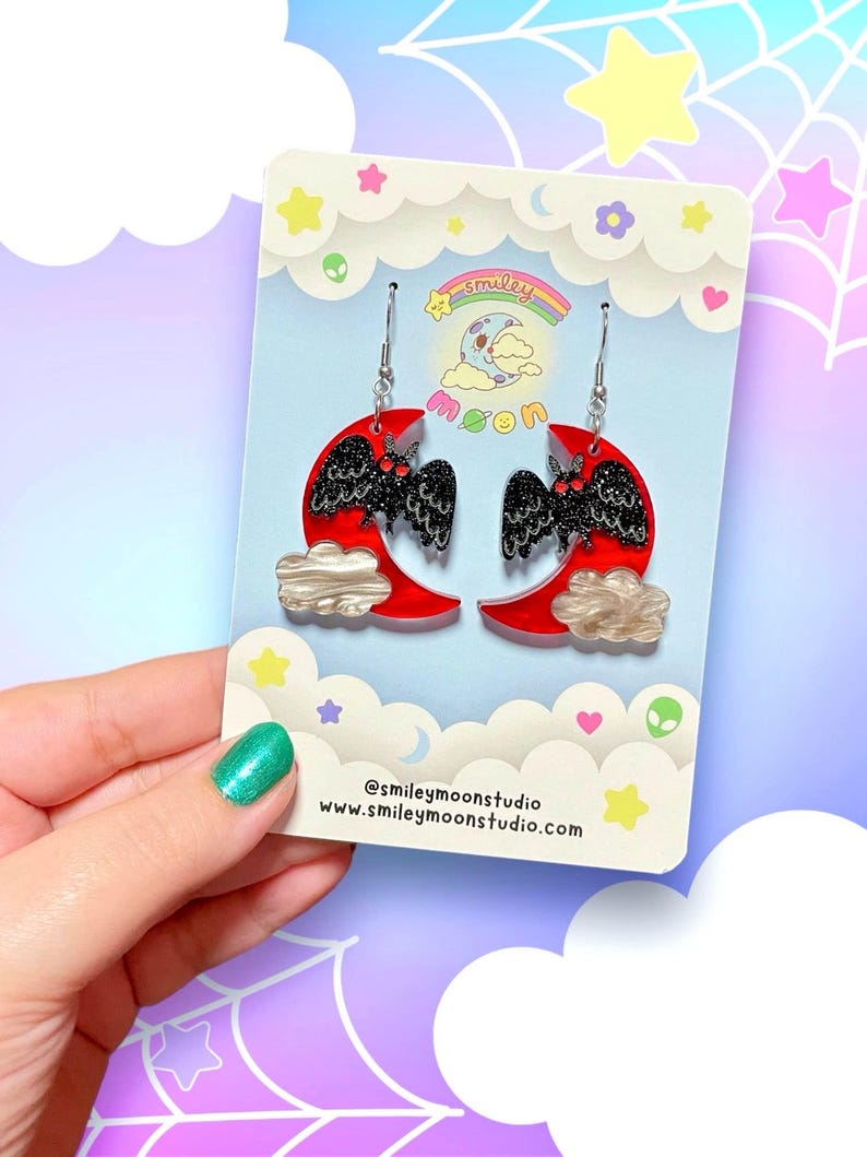 Puede incluir: Un par de pendientes de media luna roja con murci&eacute;lagos negros con purpurina y nubes blancas. Los pendientes est&aacute;n en una tarjeta con el texto "smiley moon" y "@smileymoonstudio www.smileymoonstudio.com".