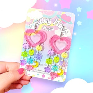 May include: Pink heart-shaped resin earrings with a dangling charm of colourful beads, stars, peace signs, smiley faces, and alien heads. The earrings are displayed on a card with the text "Smiley Moon Studio".