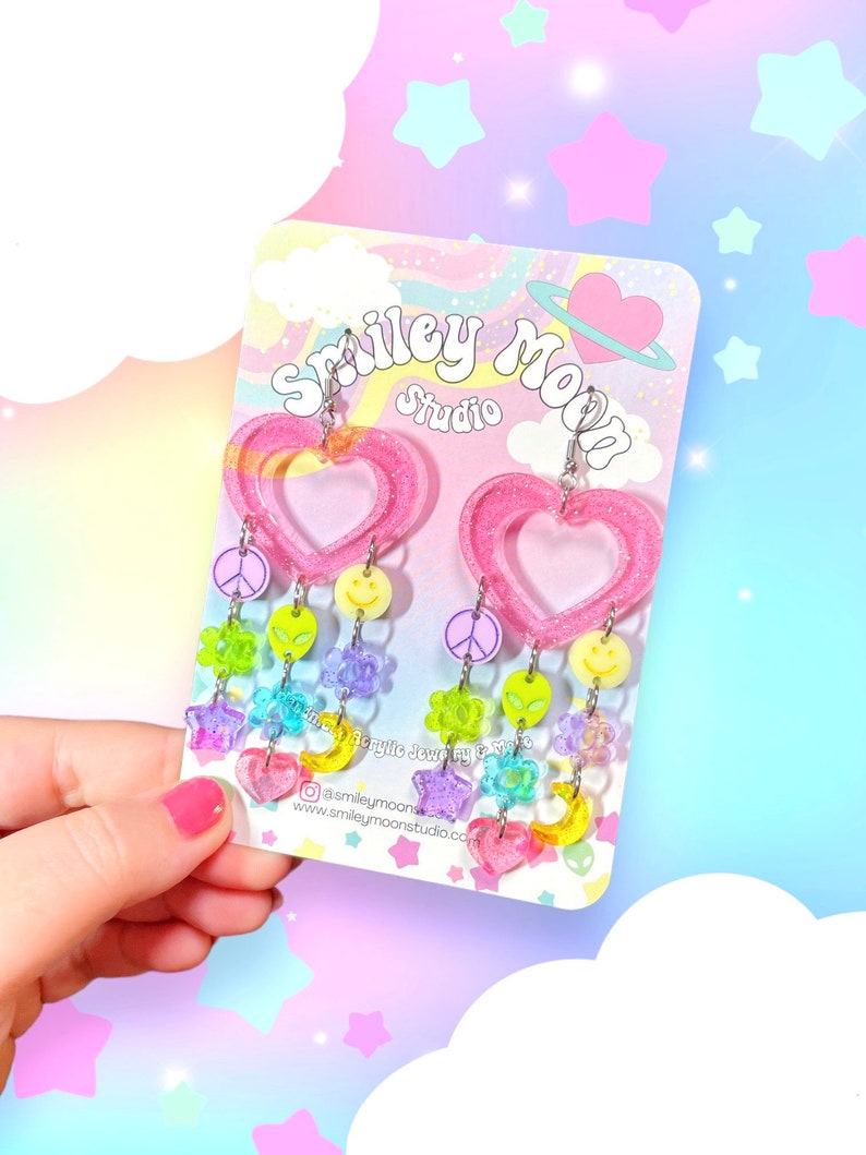 May include: Pink heart-shaped resin earrings with a dangling charm of colourful beads, stars, peace signs, smiley faces, and alien heads. The earrings are displayed on a card with the text "Smiley Moon Studio".