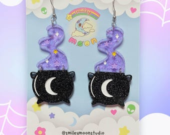 May include: A pair of purple and black glitter cauldron earrings with a crescent moon charm. The earrings are on a white card with the text "@smileymoonstudio www.smileymoonstudio.com".