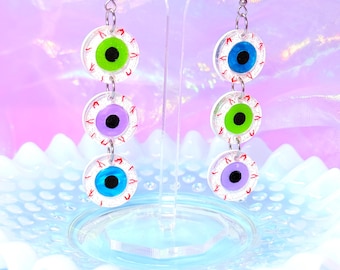 May include: A pair of dangling earrings featuring a stacked design of three clear, circular eye-shaped charms. Each charm has a colored iris (green, blue, or purple) with a black pupil and red vein detailing. The earrings hang from silver-colored hooks.