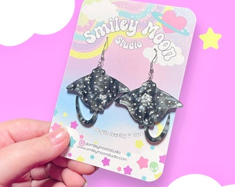 May include: A pair of black and white spotted stingray earrings with silver hooks. The earrings are on a card that says "Smiley Moon Studio" with a rainbow and a heart. The card also says "Acrylic Jewelry & More" and includes social media information.