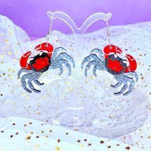 May include: A pair of crab-shaped earrings. Each earring features a red claw, a black head, and a gray body with multiple legs. The earrings are suspended from silver hooks and displayed on a clear stand against a white and gold background.