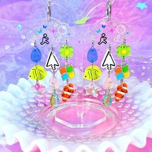 May include: Dangle earrings with a cloud-shaped top, adorned with charms including a running figure, a computer cursor, a flower, a fish, and a colourful square. The earrings are displayed on a decorative stand against a pastel background. The earrings are designed with a playful aesthetic.