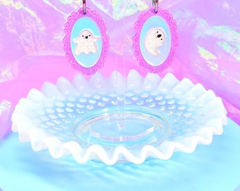 May include: A pair of ghost-themed earrings. Each earring features a light blue oval with a white ghost design, framed by a pink, ornate border. The earrings hang from silver hoops. The earrings are displayed on a clear stand, with a ruffled glass dish in the foreground.