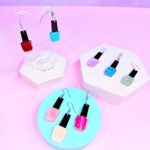 May include: Earrings shaped like nail polish bottles in various colors, including red, blue glitter, pink, and light pink. The earrings have black caps and silver hooks, displayed on white and blue platforms against a purple background.