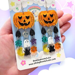 May include: A pair of dangle earrings featuring a pumpkin charm with a jack-o'-lantern face, followed by a chain of charms including skulls, stars, ghosts, moons, spiders, and flowers. The charms are in various colours, including black, white, orange, green, purple, and yellow. The earrings are on a card with the text "@smileymoonstudio.com www.smileymoonstudio.com".
