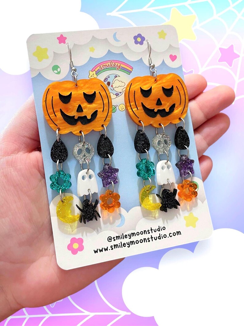 May include: A pair of dangle earrings featuring a pumpkin charm with a jack-o'-lantern face, followed by a chain of charms including skulls, stars, ghosts, moons, spiders, and flowers. The charms are in various colours, including black, white, orange, green, purple, and yellow. The earrings are on a card with the text "@smileymoonstudio.com www.smileymoonstudio.com".