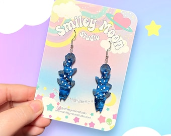 May include: A pair of blue and white acrylic earrings shaped like a fish. The earrings are on a card that says "Smiley Moon Studio" and "Handcrafted Acrylic Jewelry & Accessories".
