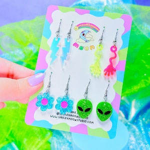 May include: A set of colorful, quirky earrings on a display card. The earrings include lightning bolts, flowers, alien heads, and abstract shapes in shades of blue, pink, and green. The card has a rainbow and the text "smiley moon".