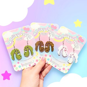 May include: Three pairs of acrylic earrings in green, gold, and white, each with a unique design. The earrings are displayed on colorful cards with the "Smiley Moon Studio" logo. The background features a pastel gradient with star and cloud accents.